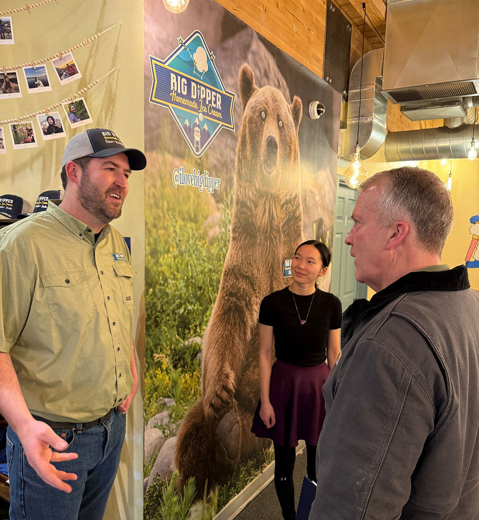Two people engage in conversation near a "Big Dipper Homemade Ice Cream" mural featuring a bear. Warm lighting and hanging photos adorn the space.