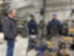 Three men in a workshop, one speaking and two listening. They are surrounded by machinery and tools. The atmosphere is focused.