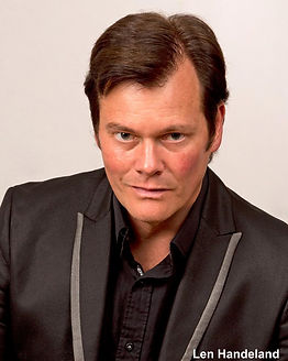 Len Handeland, award-winning author, wearing a black suit and smiling confidently.