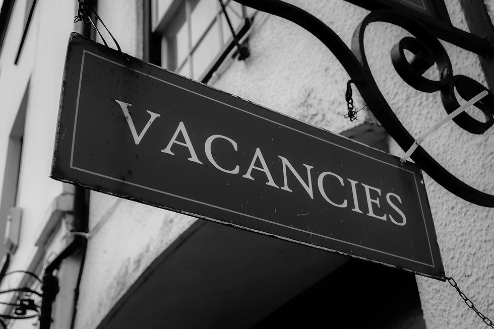 Black and white photo of a "VACANCIES" sign hanging on a building with textured walls and window in the background.