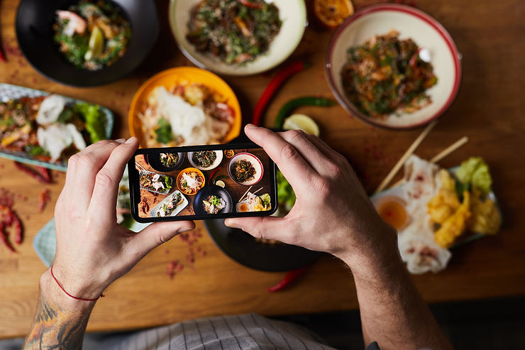 Hands holding a smartphone capturing a photo of colorful food bowls on a wooden table. Various dishes and vibrant ingredients visible. Hands holding a smartphone capturing a photo of colorful food bowls on a wooden table. Various dishes and vibrant ingredients visible.