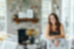 Woman smiling and standing relaxed in living room with stone fireplace, dried flowers and white furniture.