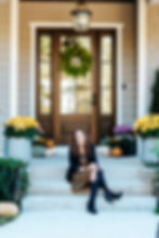 Smiling woman with brown hair sitting on front stoop surrounded by flowers and pumpkins.