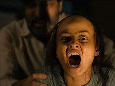A girl in a dim setting is screaming with her mouth wide open, displaying fear or shock. A person in the background holds her shoulders.