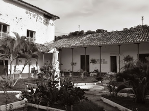 Patio interior de una casa antigua con techo de tejas, plantas y una estatua en el centro. Ambiente tranquilo y monócromo.