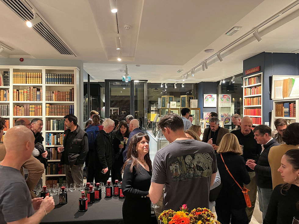 People socialize in a bookstore with shelves of books. Bottles and glasses are on a table. The mood is lively with warm lighting.