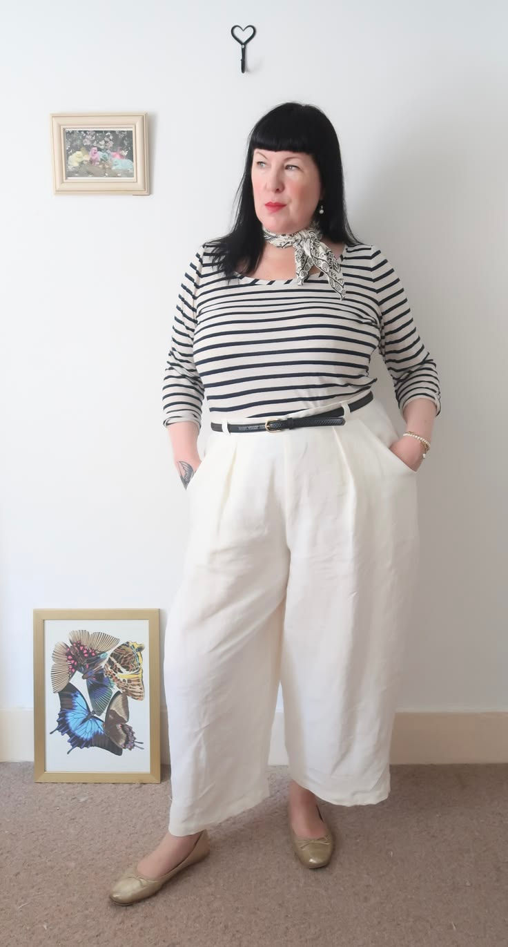 Woman in a striped top and white pants stands indoors beside framed art, hands in pockets, looking off to the side.