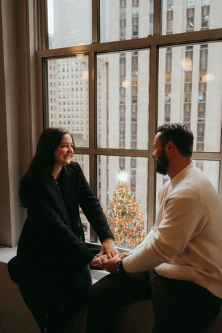 Romantic couple photoshoot by window Rockefeller Tree Christmas in NYC holding hands smiling with engagement ring