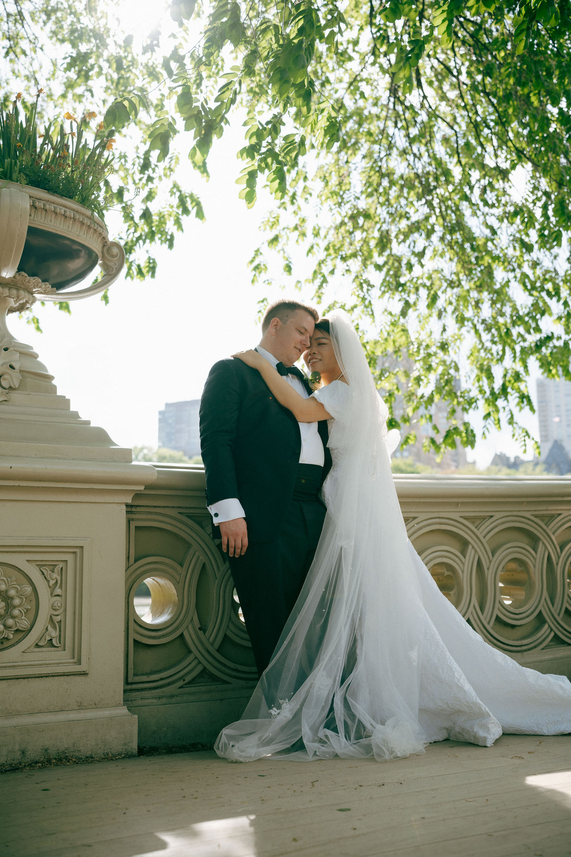 Central Park wedding elopement photography with bride in white gown and groom in black suit embracing on Bow Bridge surrounded by leafy trees during daytime
