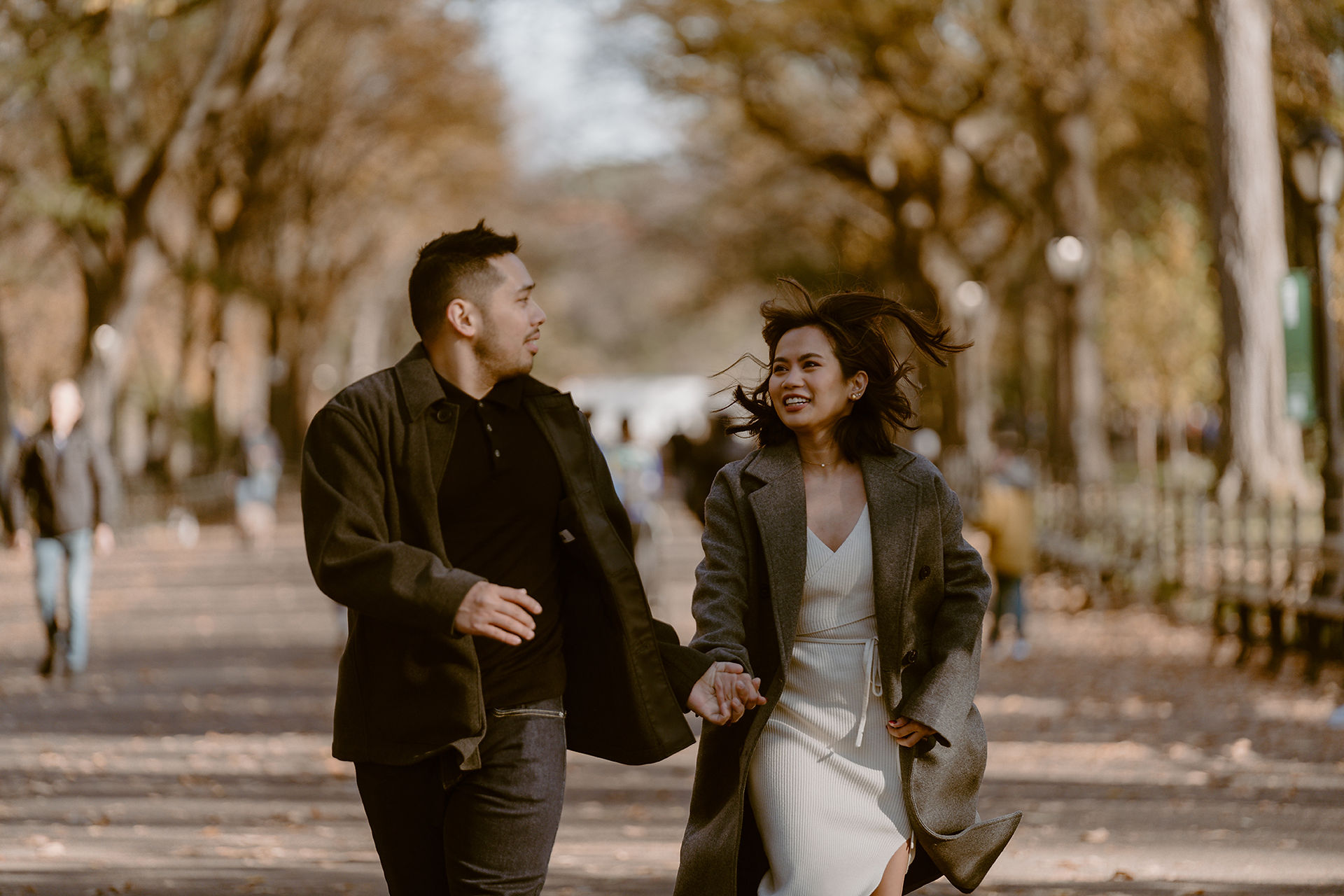 A couple joyfully runs hand in hand down a tree-lined pathway at Bethesda Terrace in Central Park, NYC, during their romantic fall photoshoot. The golden autumn leaves add a cinematic aura to this portrait session.