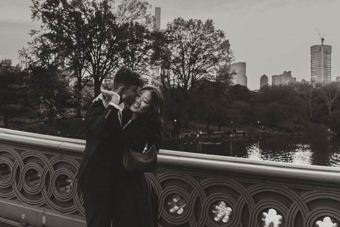 Romantic couple portrait on Bow Bridge, NYC with city skyline and trees in cinematic black and white.