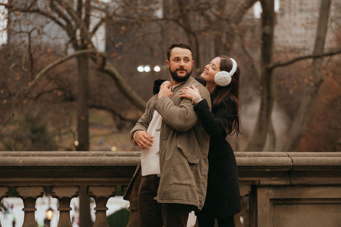 Couple hug in Central Park during marriage proposal photoshoot in New York, cityscape backdrop, relaxed and content, romantic photoshoot engagement at 620 loft and garden proposal