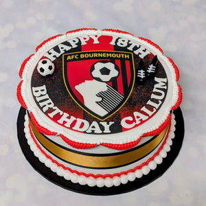 White iced cake on a black board with white and red piping around top and bottom edge. A gold with black edge ribbon around cake. A Bournemouth Football badge edible print covers top of cake with plastic football and pair of small football boots.