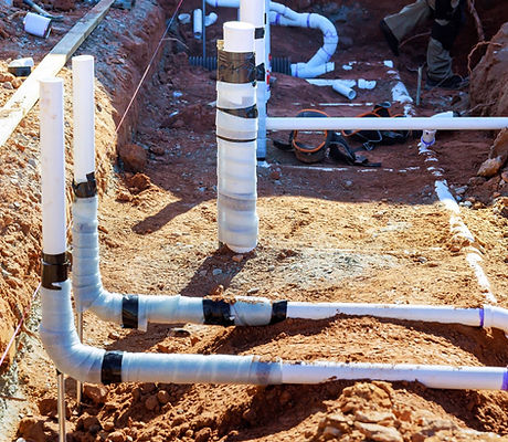 A network of white PVC drain and sewer pipes laid out in an excavated trench with red clay soil