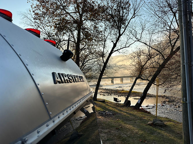 AIRSTREAM trailer by river, trees, and distant bridge