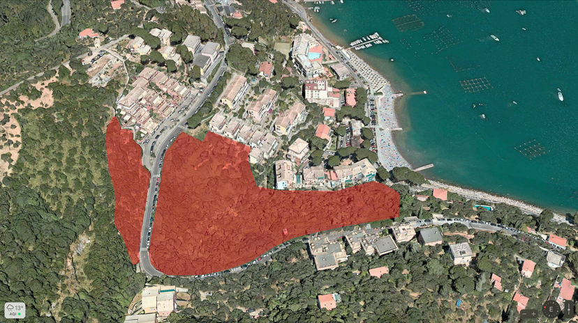A satellite view of a coastal area with a sandy beach along the shoreline, surrounded by lush greenery and residential buildings. A large red highlighted area marks a section of forested land inland, with the deep blue sea on one side and a road running parallel to the beach on the other.