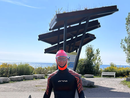 Coach Robert in black wetsuit and pink helmet stands by an orange kayak at a lakeside park under a modern shelter.