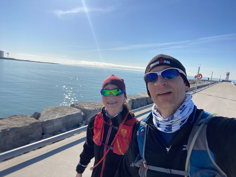 Two people in sunglasses and running gear smile by a waterfront path on a sunny day. Blue sky and calm water in the background.