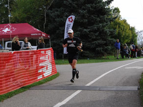 Coach Robert crosses finish line on a park path, cheered by spectators by a medical tent. Red barriers and trees in the backdrop.