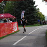 Coach Robert crosses finish line on a park path, cheered by spectators by a medical tent. Red barriers and trees in the backdrop.