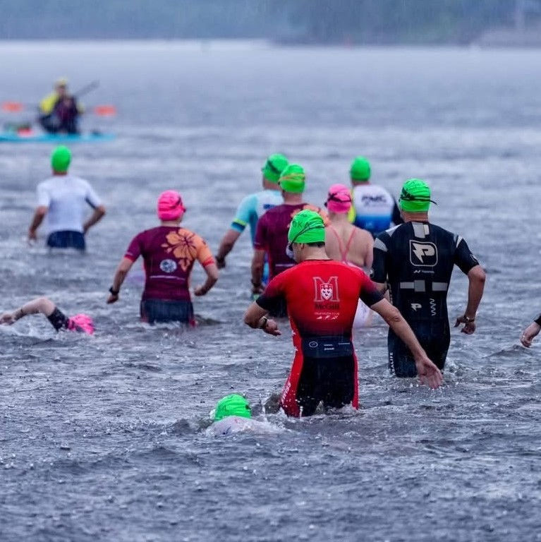 Swimmers in neon caps wade through choppy lake water during an open-water race, with a kayak nearby and a misty shoreline.