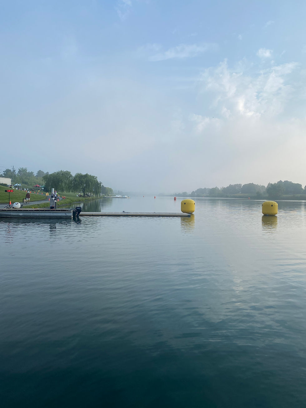 Triathlon Swim Start with calm water, yellow buoys, a dock, and a few people on the shore under a pale blue sky.