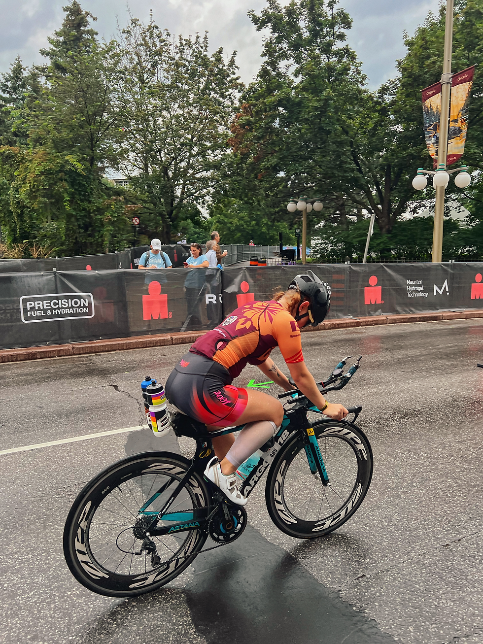 Cyclist in orange-and-maroon racing gear rides a triathlon bike on a wet road, with sponsor banners and trees behind her.