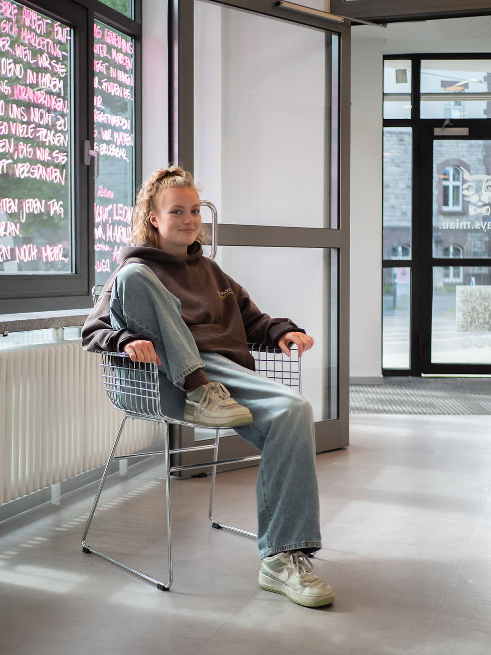 Junge Frau sitzt entspannt in einem Drahtstuhl in einem modernen Bürogebäude. Sie trägt Hoodie, Jeans und helle Sneaker. Neben ihr ein großes Fenster mit pink beschrifteten Tafeln.