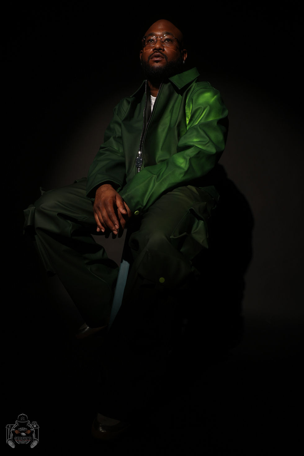 Man in green jumpsuit sitting, illuminated by studio light, dark background.