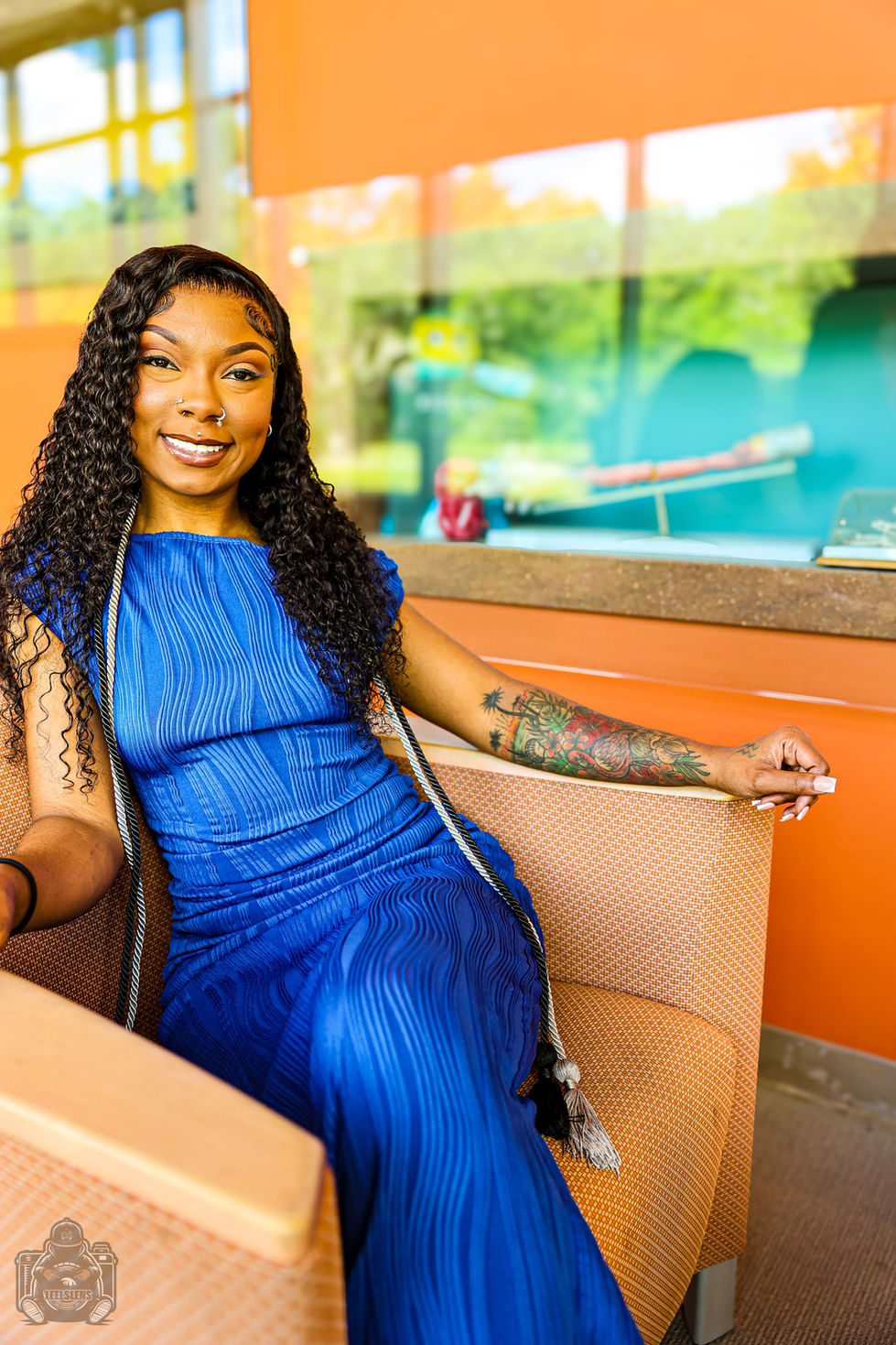 Smiling Black woman in blue pleated dress, long curly hair, arm tattoo.