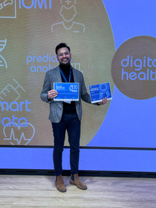 A man in a beige blazer, black turtleneck, and dark pants stands smiling on stage, holding two certificates. Behind him, a large digital screen displays "digital health" and various medical and technology-related terms.