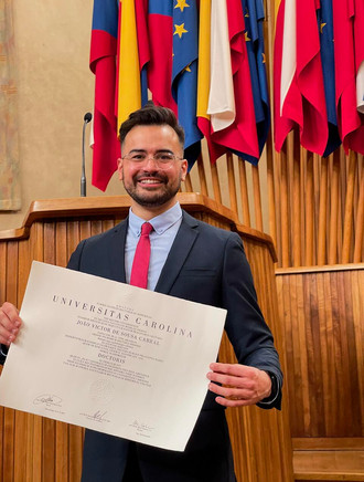 Joao Victor Cabral holding his official doctoral diploma at Charles University, standing in front of an elegant wooden podium adorned with national and European flags.
