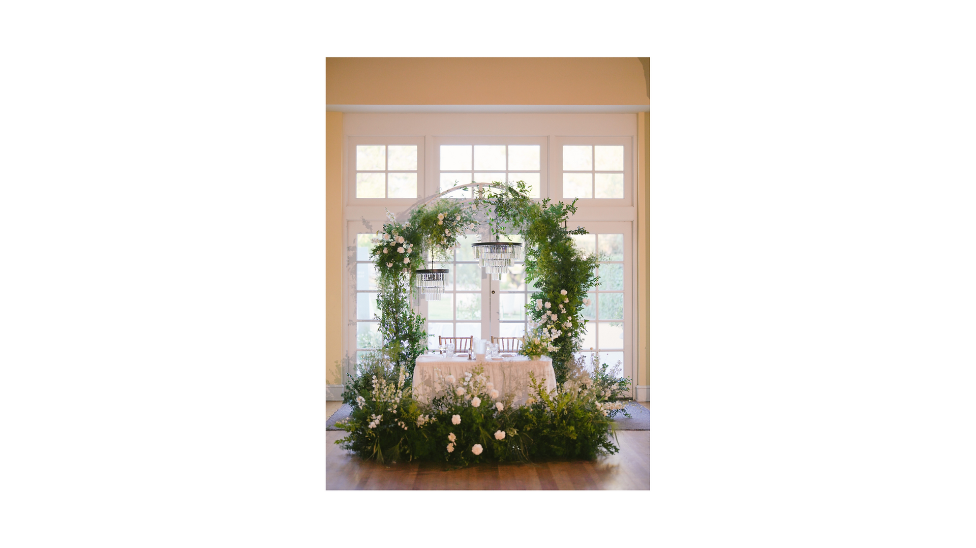 Real wedding sweetheart table featuring a floral-adorned arch, crystal chandeliers, a white draped table, and lush greenery, designed by Ido Events for a luxurious and romantic ambiance.