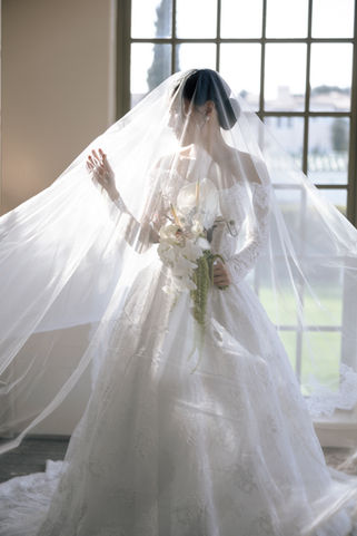 Bride in lace gown holding white floral bouquet under sheer veil, standing in front of a large window at Marbella Country Club, styled by IDO Events.