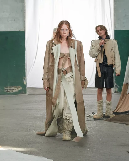 A model walks the runway at Deadwood’s SS24 collection during Copenhagen Fashion Week in a raw, industrial setting. She is wearing a floor-length, distressed brown trenchcoat handcrafted in Sweden from upcycled Volvo leather offcuts. The outfit features a minimalist strap-style top and beige utility trousers, paired with rugged tactical boots. In the background, a person stands in a cropped tan jacket and black leather shorts against white draped fabric, highlighting the brand's focus on sustainable, high-end circular design.
