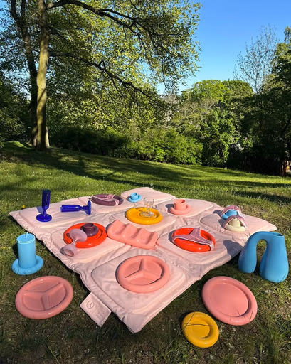 An outdoor installation featuring a pink padded picnic mat and colorful, sculptural tableware with Mercedes-Benz branding. Set in a green park environment, showcasing a technical textile collaboration produced by XV Production.
