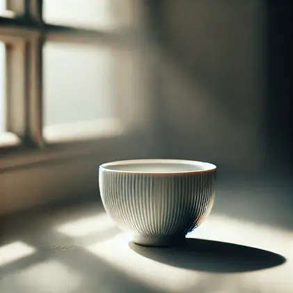 A white, ribbed bowl sits on a sunlit table by a window, casting a shadow. The soft light creates a calm, serene atmosphere.