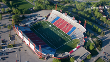 Autonomous security drone conducting aerial surveillance over a stadium and arena complex during a major sporting event, monitoring the parking lot, crowd approach corridors, and perimeter from above