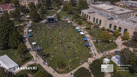 Aerial view of a large campus event with crowds gathered on a central lawn, stage setup visible, pathways filled with people, and surrounding university buildings.