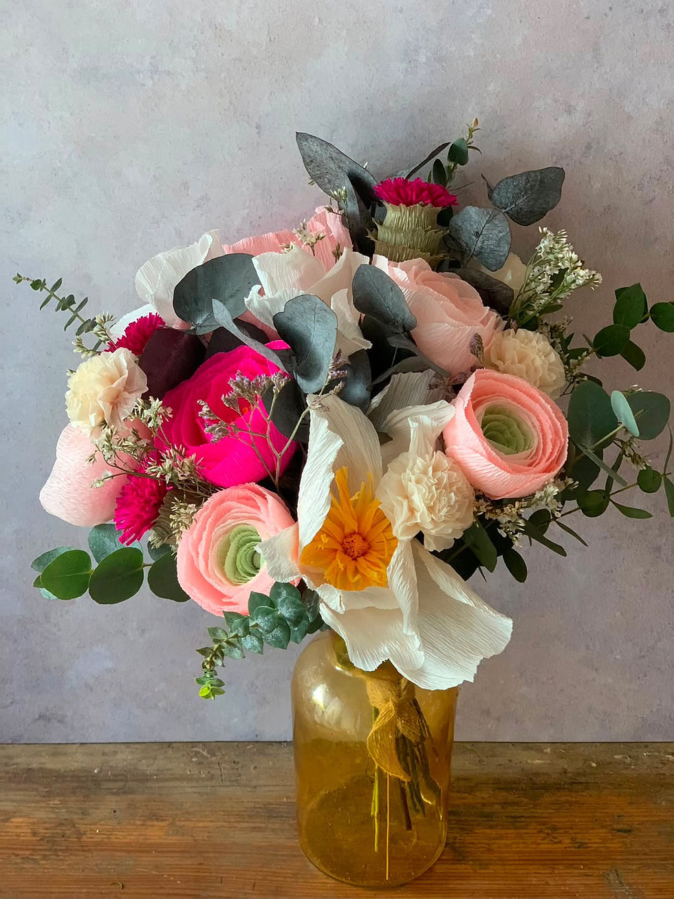 Colourful bouquet of pink, white, and yellow paper flowers with paper eucalyptus in an amber glass vase on a wooden table.