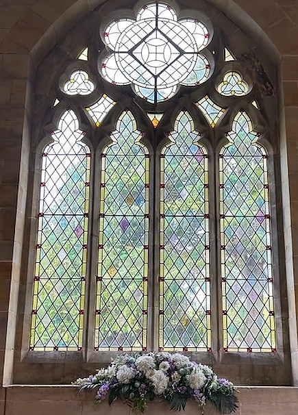 A beautiful stained glass arched window in a funeral chapel with an arrangement of flowers underneath