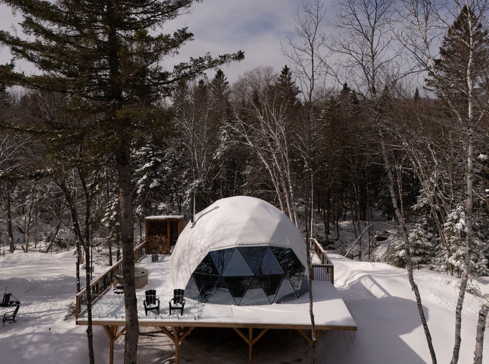 Vue aérienne du Dôme L'Épinette en pleine forêt enneigée, avec grande terrasse privée, spa à jets et espace foyer extérieur.