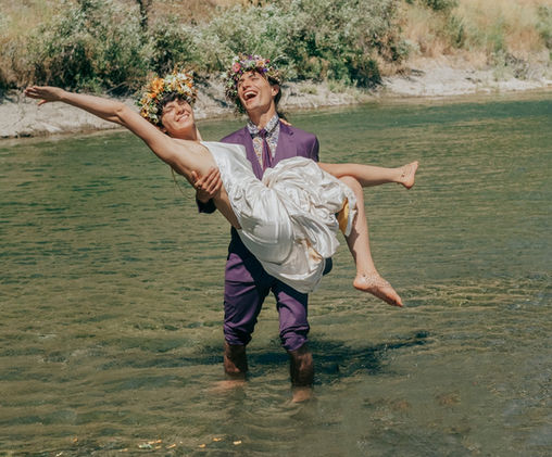 Man carrying woman in water, both wearing flower crowns, laughing merrily.