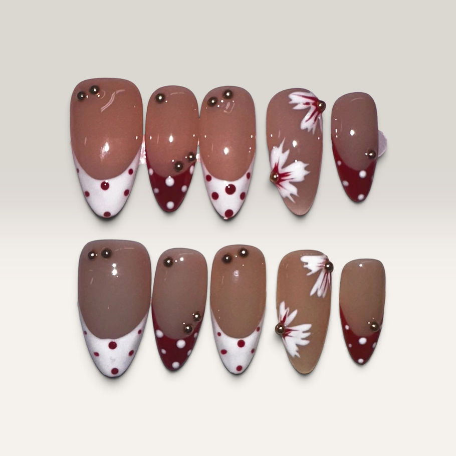 Cherry Blossom Chic press-on nails featuring a glossy nude base with burgundy and white polka-dot French tips, delicate white floral accents, and small gold bead embellishments in an elegant almond shape.