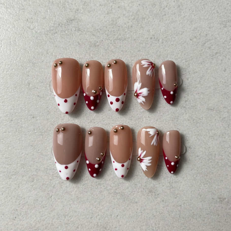 Cherry Blossom Chic press-on nails featuring a glossy nude base with burgundy and white polka-dot French tips, delicate white floral accents, and small gold bead embellishments in an elegant almond shape.