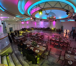 El Salón Casino Real es un espacio espectacular y de gran capacidad, diseñado para eventos festivos y recepciones a gran escala. Su diseño interior es moderno, con pisos de mármol y un techo de diseño circular que integra iluminación LED y una bola disco central, creando un ambiente de club nocturno elegante. Cuenta con un escenario amplio, dos grandes pantallas de proyección y una pista de baile central. La ambientación utiliza mesas rectangulares de madera oscura y sillas Crossback, lo que lo hace perfecto para bodas, XV años y eventos corporativos que requieran un ambiente vibrante y totalmente equipado.