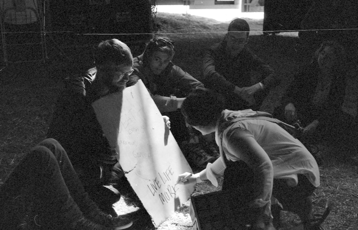 Image Description: A black and white photoof a group of people sitting outside, Kerri writes 'LIVE LIFE MORE' on a board that is held up by another member of the group.