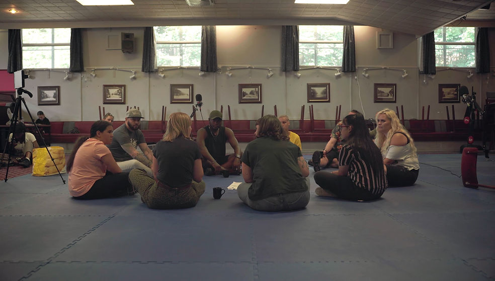 A group of people of various ages sit on the floor in a circle in the hall at a working men's club talking. The floor has soft judo matts on it, windows let sun in and there are framed black and white pictures on the walls