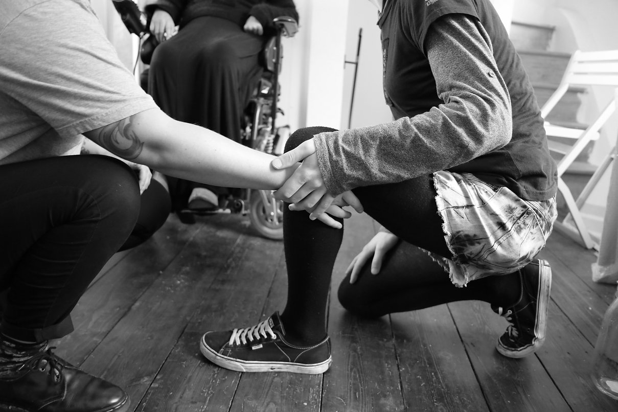 Image Description: A black and white image of two people crouching, one holding the other's knee whilst the other holds their arm. A person in a wheelchair sits in the background.