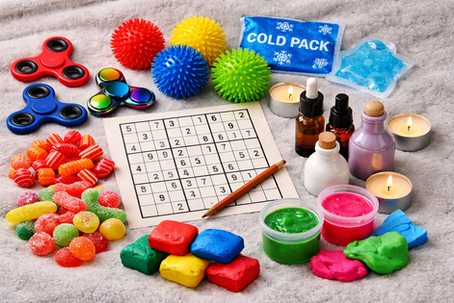 Verschiedene Skills und Sinnesmaterialien liegen auf einem weichen Untergrund ausgebreitet, darunter Fidgetspinner, Igelbälle, saure und scharfe Bonbons, Cold Packs, Duftöle, Knete, Slime und ein Sudoku mit Stift, alles übersichtlich und farbenfroh arrangiert.